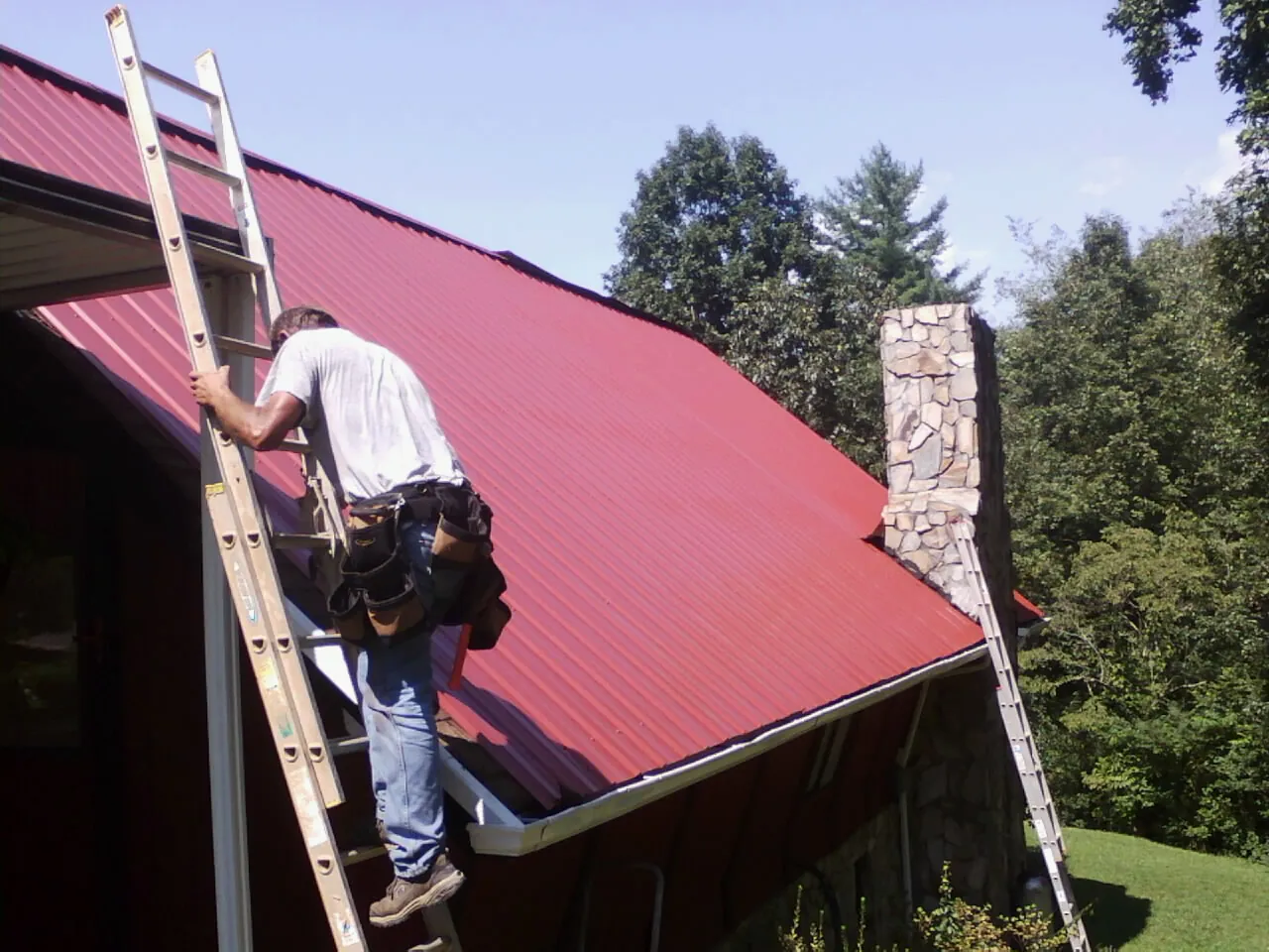 Roofer on a ladder beside a red metal roof and stone chimney — Soffit & Fascia Repair by Roofing Brothers in Yellow Spring